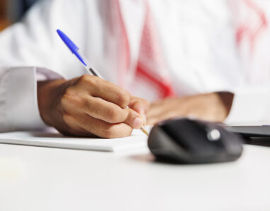 Middle Eastern freelancer efficiently works at his desk, scribbling in his notebook. He conducts online research, takes notes, and stays connected using wireless technology.